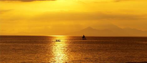 Atardecer sobre un mar tranquilo con un velero y una montaña a lo lejos.
