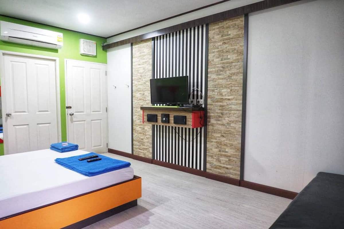 A hotel room with a bed, a TV mounted on a wooden panel wall, and a small shelf with electronic devices.