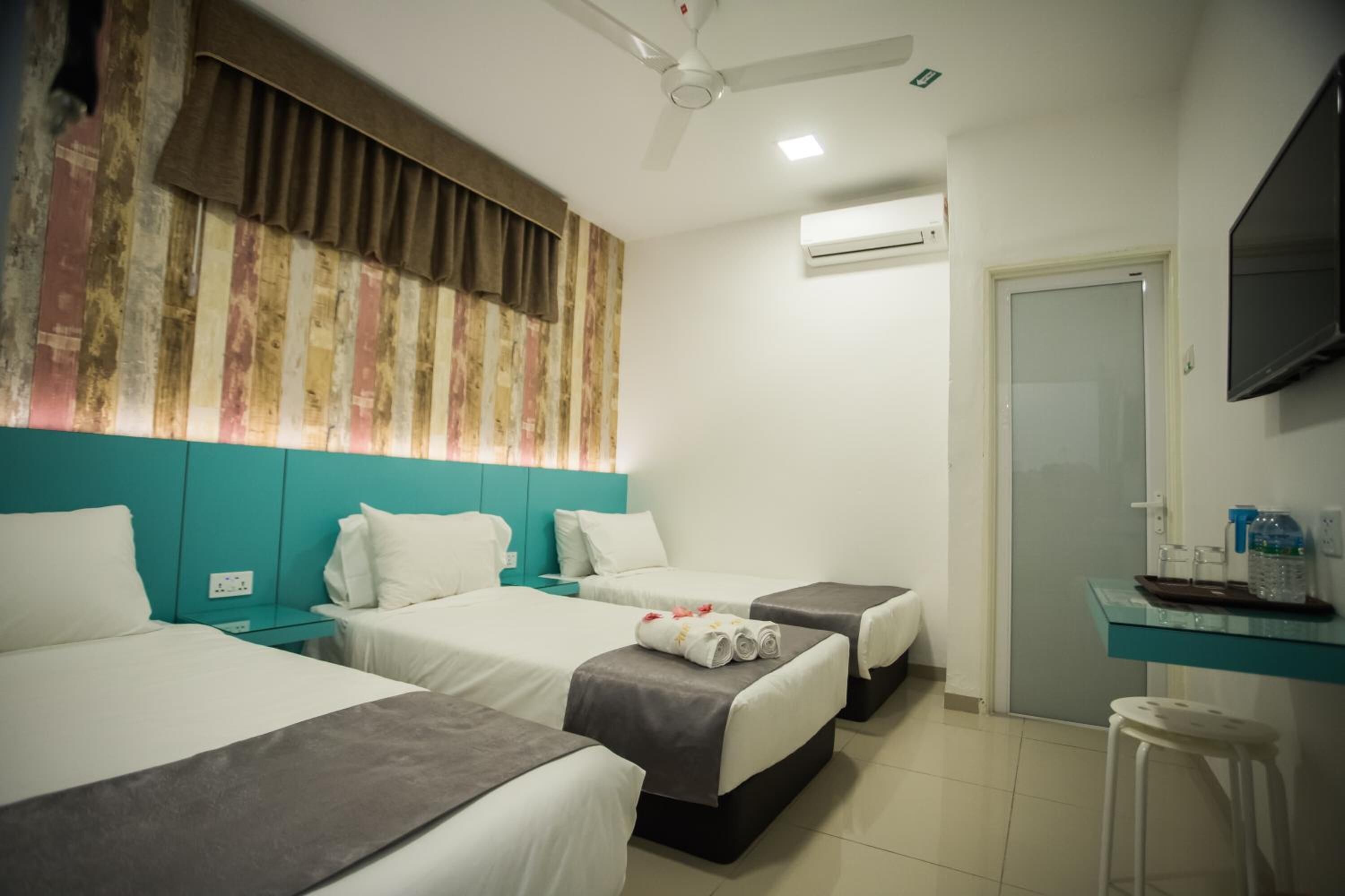Habitación de hotel con tres camas, ventilador de techo, aire acondicionado, televisión y una mesita con una botella de agua.
