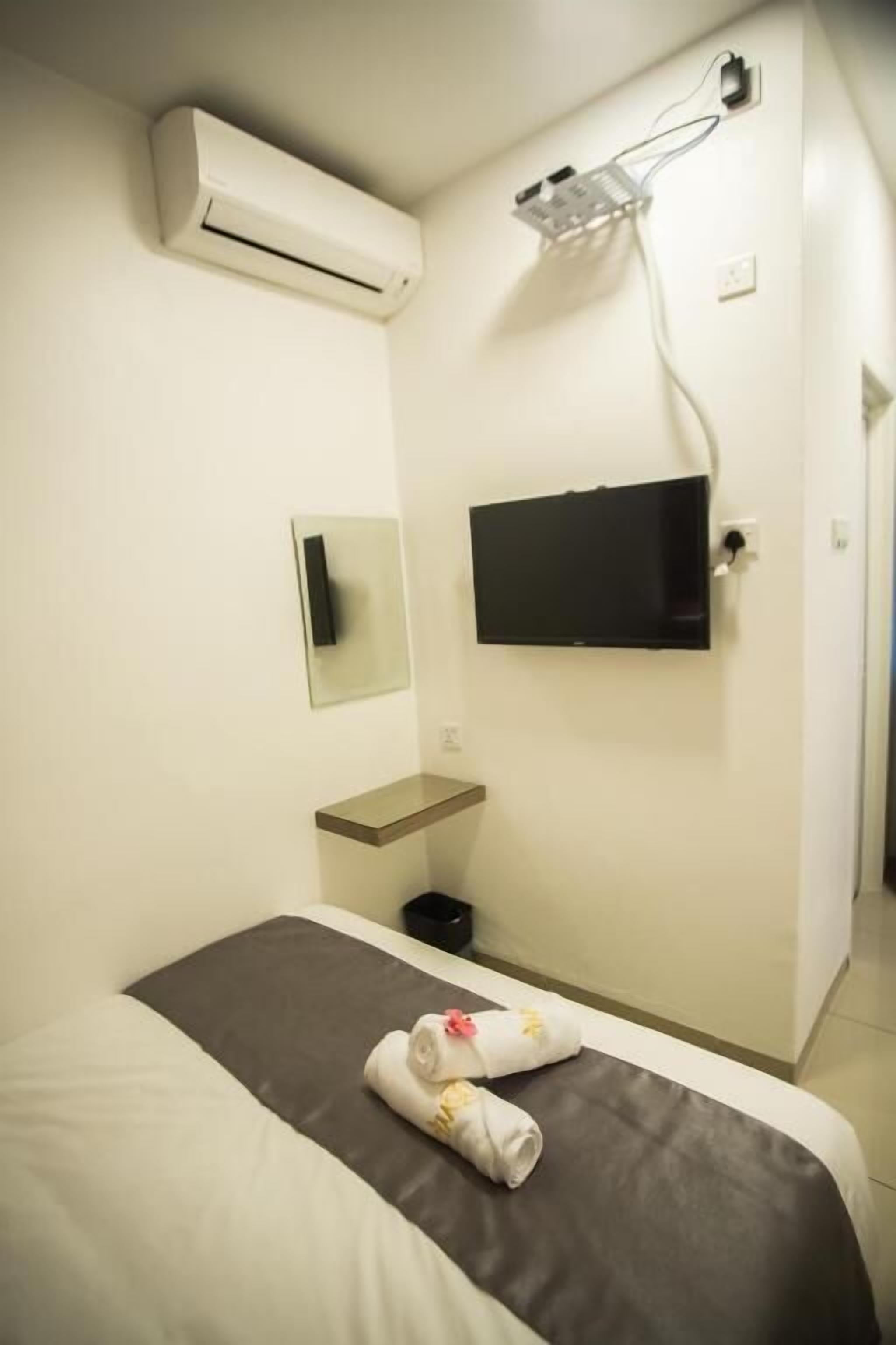 A hotel room with a bed, a flat-screen TV mounted on the wall, an air conditioning unit above the bed, a small shelf, and a towel on the bed.