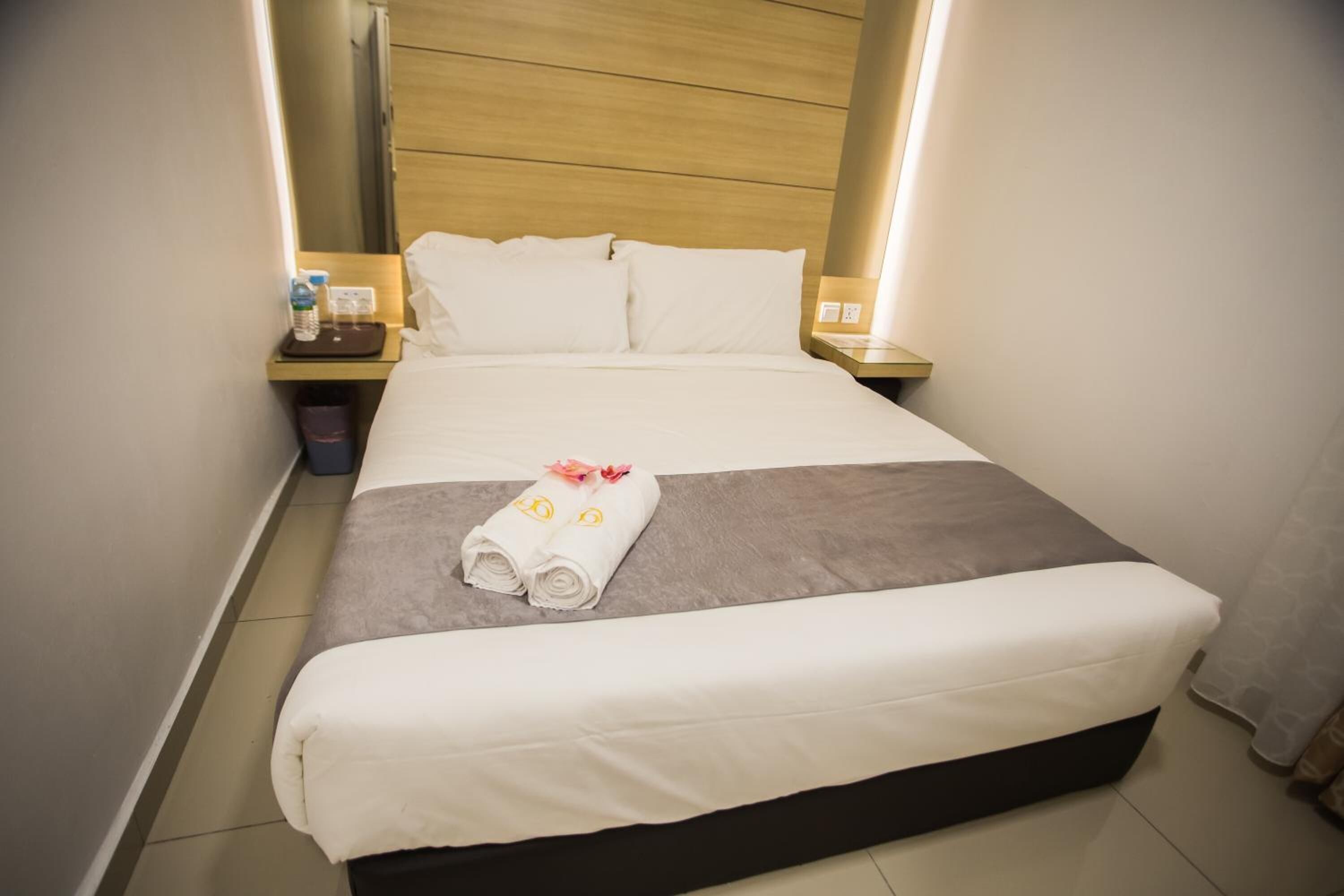 A neatly made bed with white and gray linens, a folded towel with a floral design, and a small table with a water bottle and a tissue box.