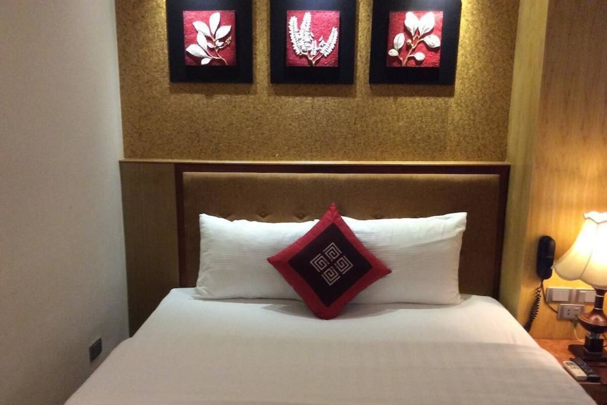 Habitación de hotel con cama, cabecera, mesita de noche con lámpara y tres cuadros con motivos florales en la pared.