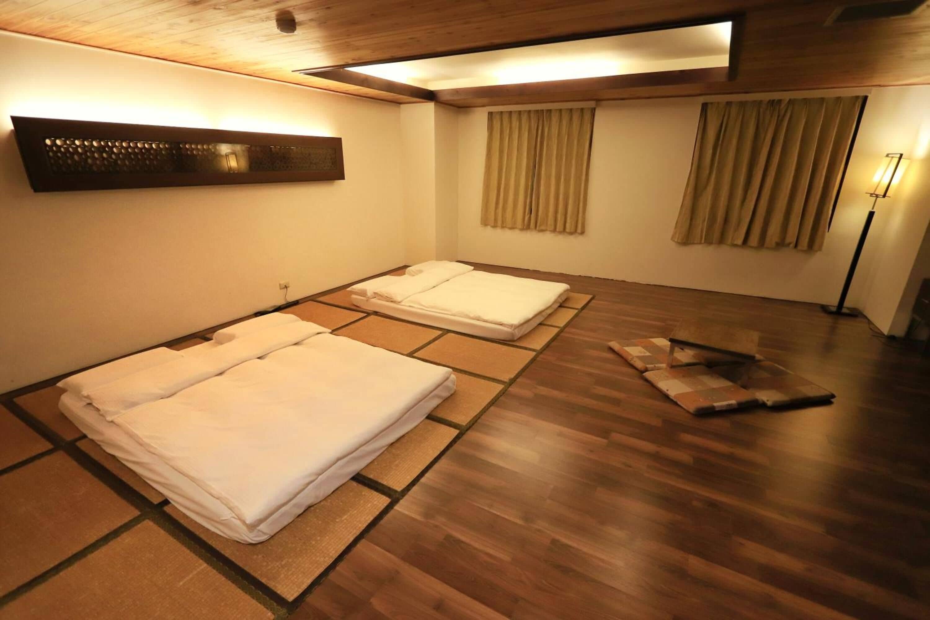 Una habitación con dos camas, suelo de madera, una lámpara de pared y una lámpara de pie.