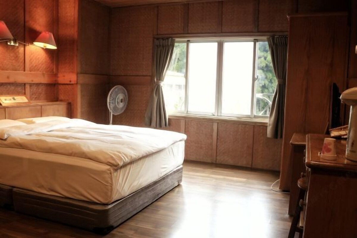 Une chambre à coucher avec un lit, une fenêtre avec des rideaux, un ventilateur sur pied et un lambris en bois.