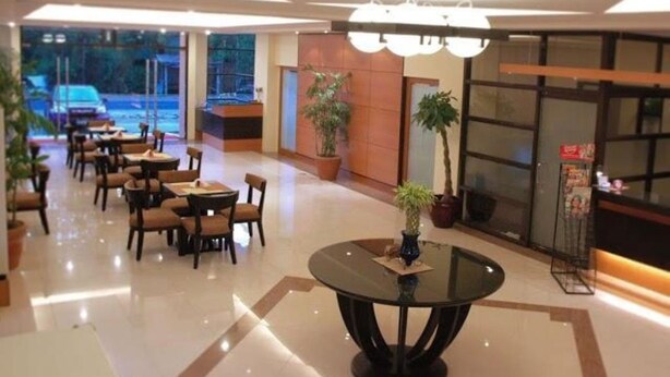 Un vestíbulo de hotel con una mesa redonda de vidrio, varias mesas de comedor, sillas, plantas en macetas y un ventanal grande.