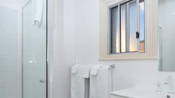 Ein Badezimmer mit Duschkabine aus Glas, einem weißen Waschbecken und einem Fenster über dem Spiegel.