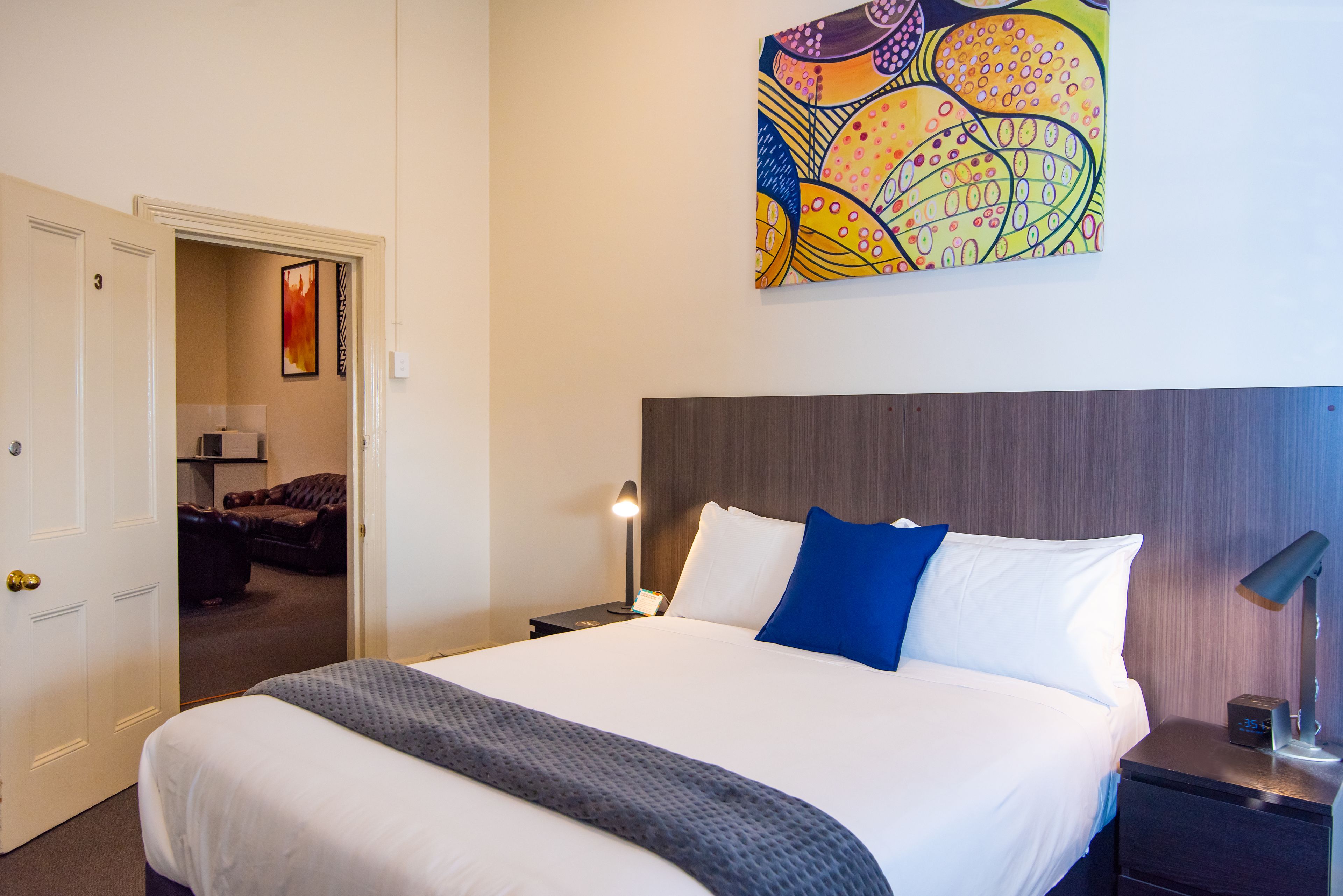 Habitación de hotel con una cama grande, una mesita de noche con una lámpara, un cuadro en la pared y una puerta que da a otra habitación.