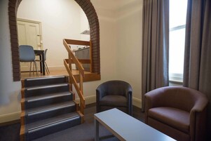 A living room with a staircase, a sofa, a table, and a view of a kitchen and dining area.