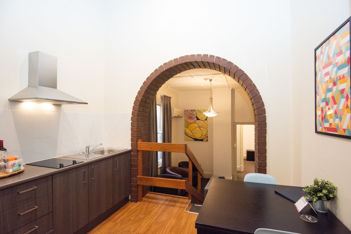 A compact kitchen with dark wooden cabinets, a stainless steel range hood, and a wooden bench at the entrance.