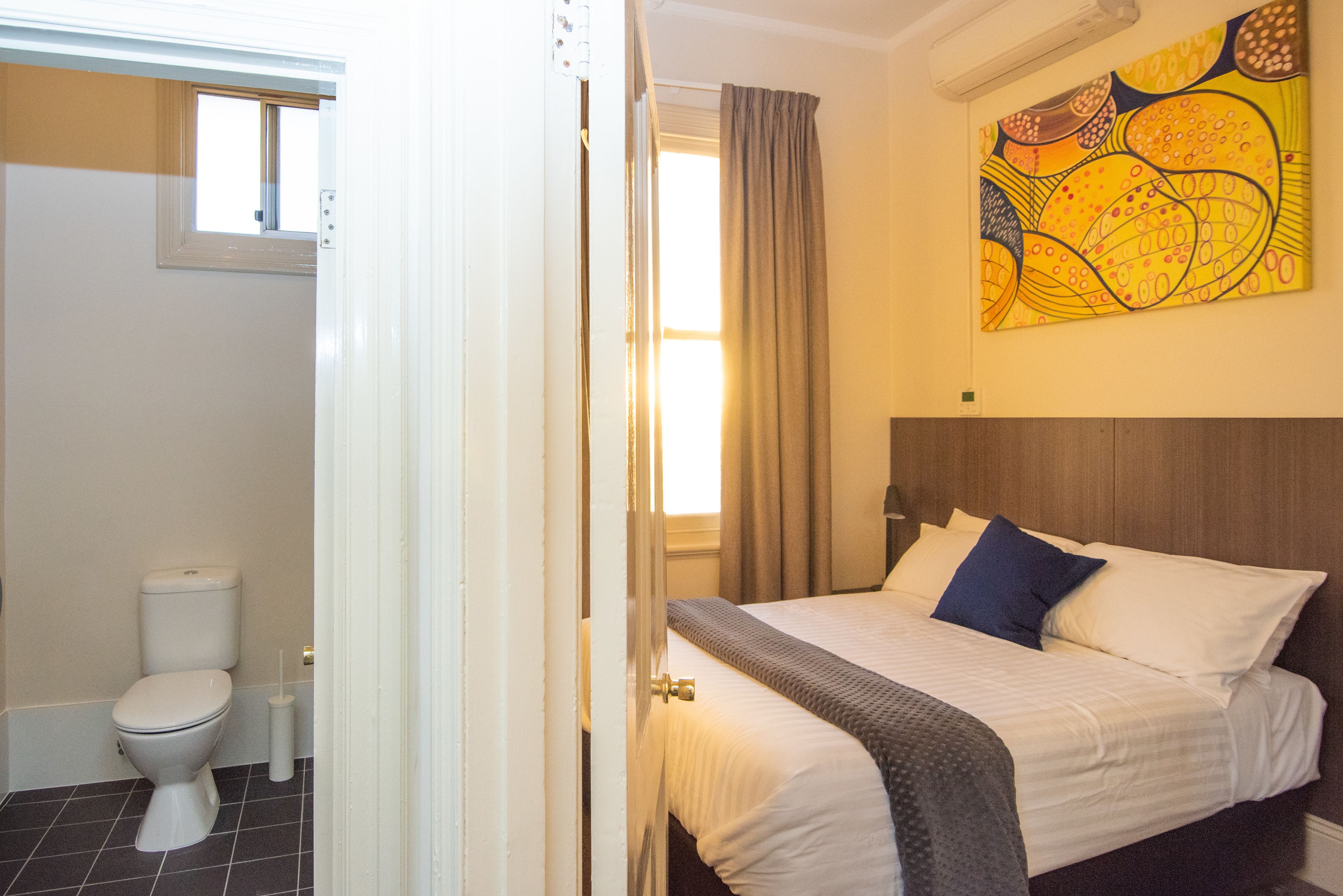 A hotel room with a bed, a window, a painting on the wall, and a bathroom with a toilet.