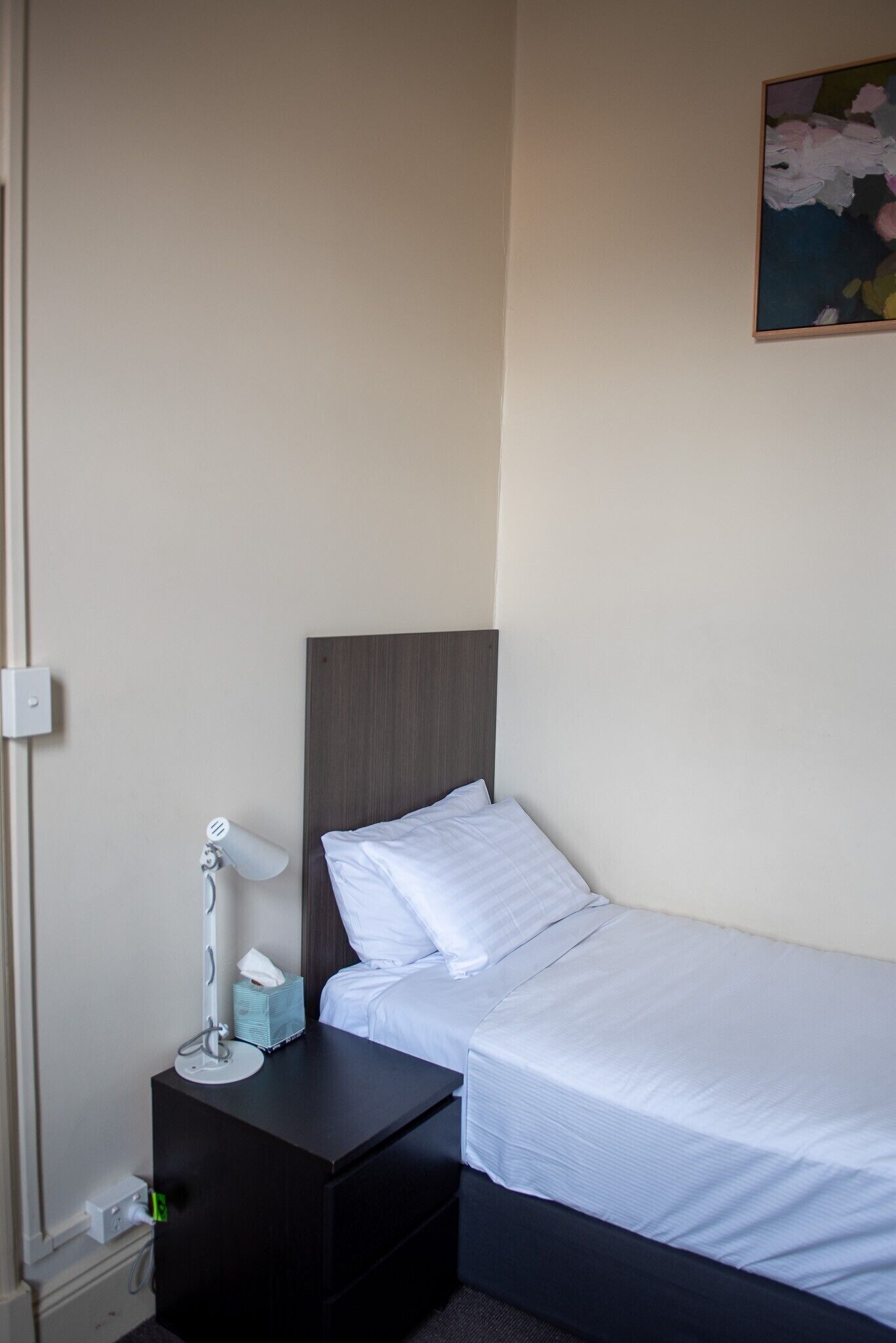 A small, neatly made bed with white linens, a dark headboard, and a bedside table with a lamp and a tissue box.