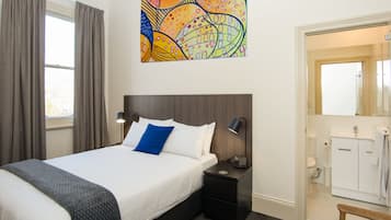 Habitació d'hotel amb un llit, taules de nit, una làmpada de nit, un bany amb un rentapins i un vàter, i un quadre abstracte i colorit a la paret.
