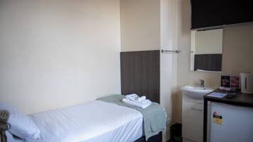 Habitación de hotel con una cama individual, una mesita de noche pequeña con una lámpara, un lavamanos y un espejo.