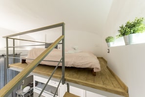 Een stapelbed met houten frame en metalen trap, een klein bureau met stoel, en een potplant op een plank.