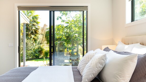 Een slaapkamer met een bed, kussens en uitzicht op een tuin door een schuifdeur.