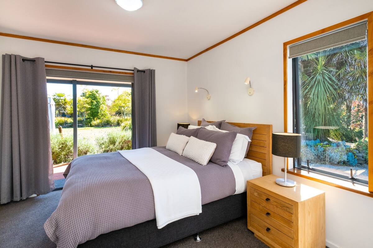 A bedroom with a bed, bedside table, lamp, and a window with a view of greenery.