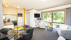 Een moderne keuken met een magnetron, oven en spoelbak. Er is een eethoek met een glazen tafel en stoelen. In de woonkamer staat een bank en een televisie.