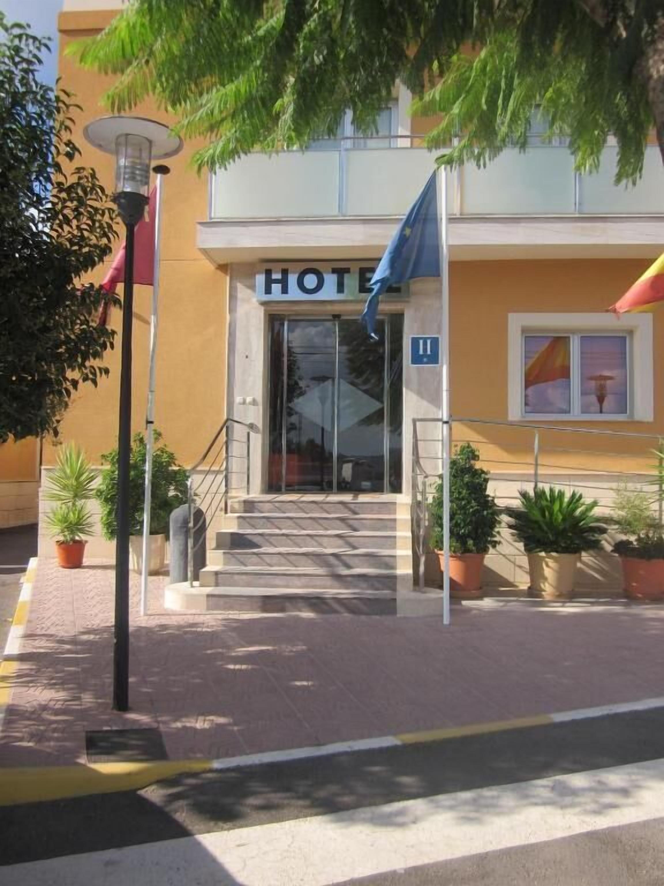 A hotel entrance with a sign reading "HOTEL" and a blue flag. There are steps leading up to the entrance, potted plants, and a street lamp.