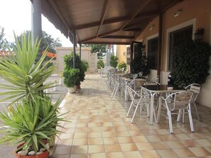 Un espace extérieur abrité, aménagé avec des chaises et des tables en métal, des plantes en pot et un sol carrelé.