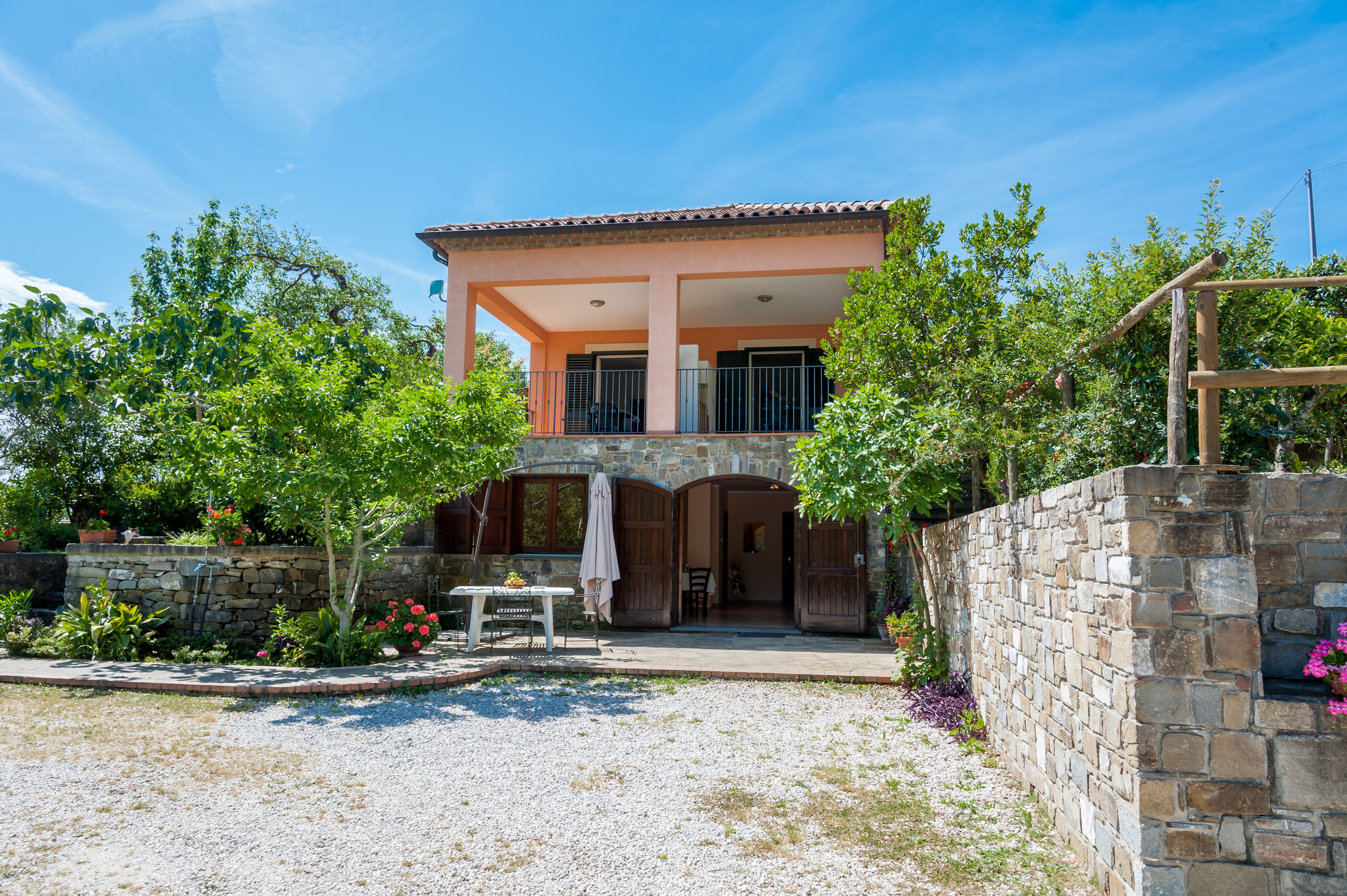 Ein zweistöckiges Haus mit einer Steinmauer, einer Terrasse mit Tisch und Stühlen und einem Garten mit verschiedenen Pflanzen und Blumen.