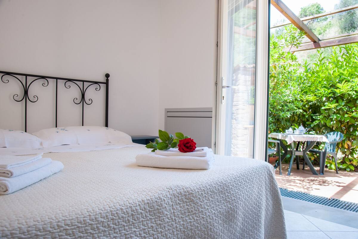 A neatly made bed with white linens and a red rose, a small table with a white tablecloth, and a view of greenery through a glass door.