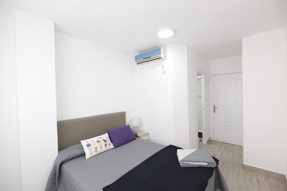Een slaapkamer met een bed, nachtkastje, een wandairconditioner en een deur.