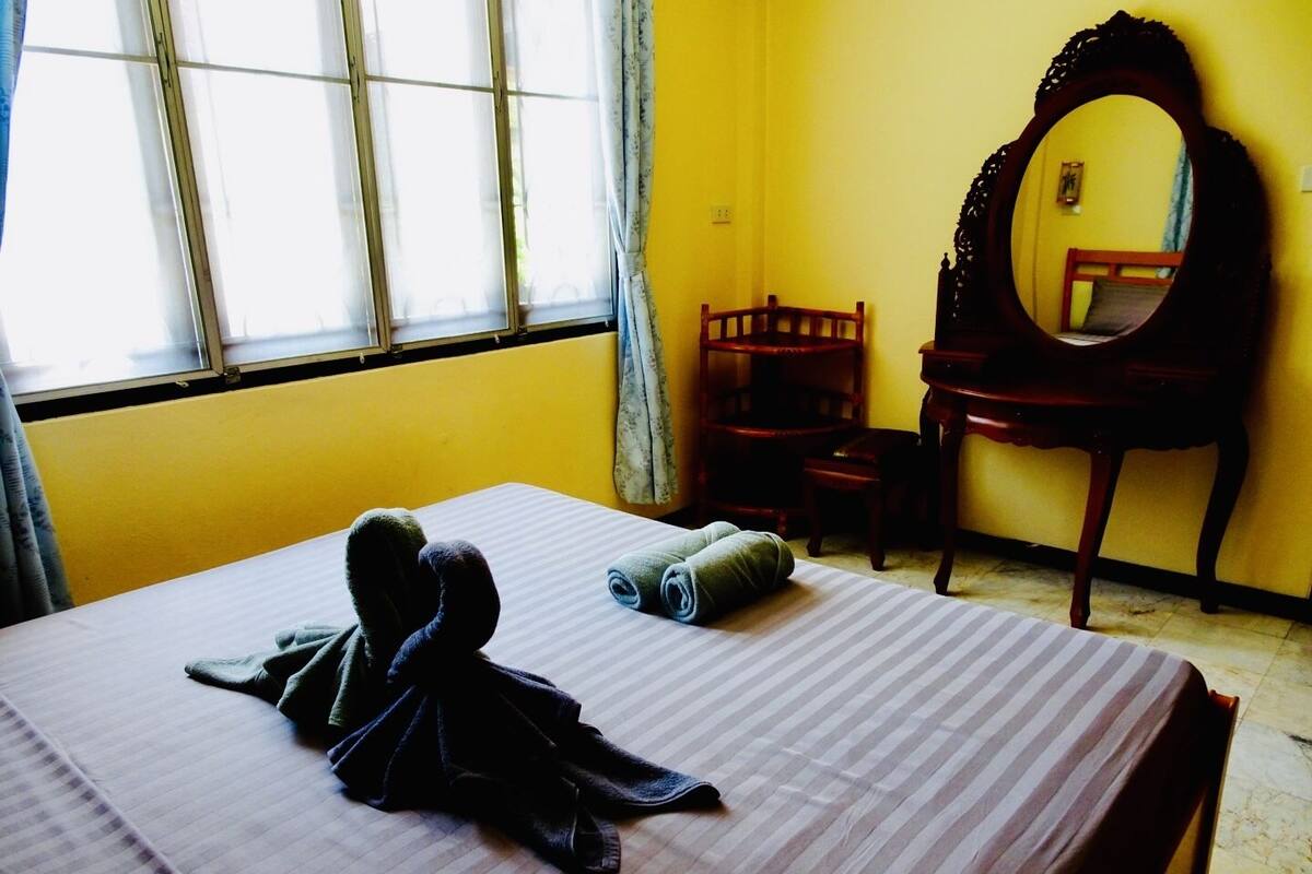 A room with a bed, a chair, a wooden table, a mirror, and a window with curtains.