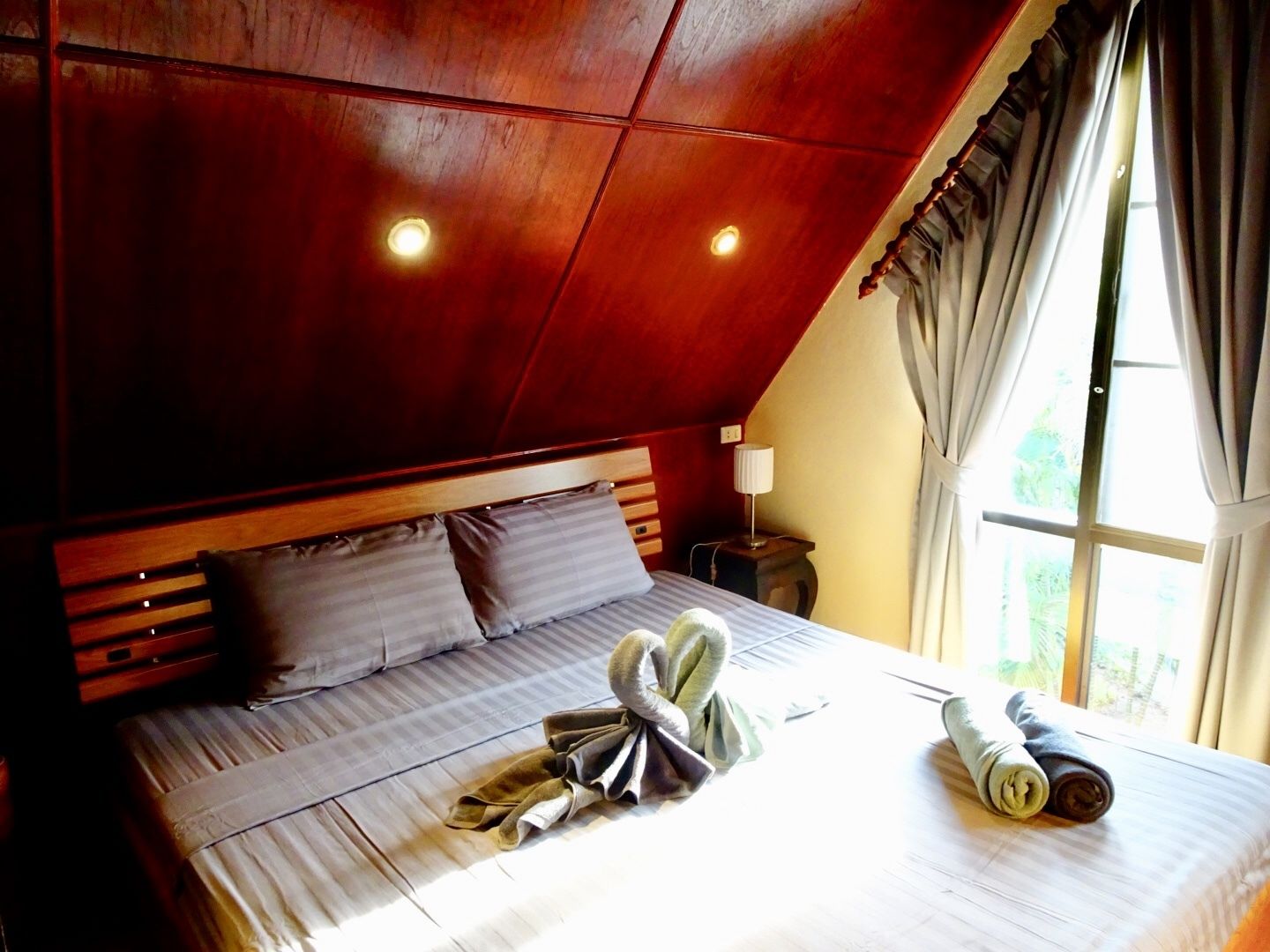 Una camera da letto con soffitto a falde, una testiera in legno, un letto con biancheria grigia, due cuscini, una lampada da comodino e una finestra con tende.