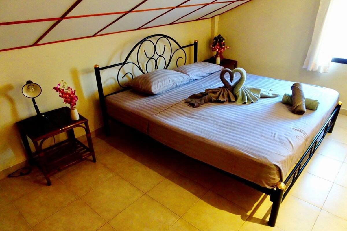 A neatly made bed with a heart-shaped towel arrangement, a bedside table with a lamp, and a vase with flowers.