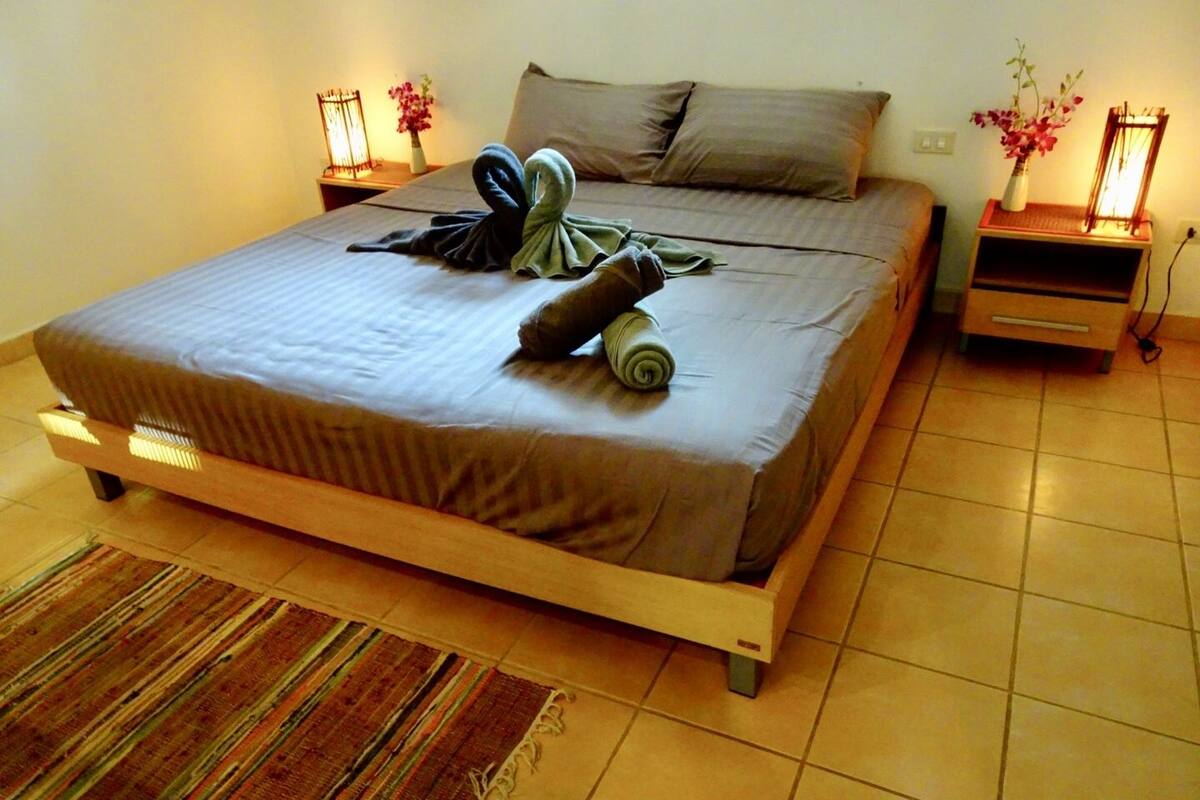A neatly made bed with a brown mattress, a striped rug, and a small wooden nightstand with a lamp and a vase of flowers.