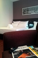 A hotel room with a bed, pillows, a remote control, and a magazine on a table.
