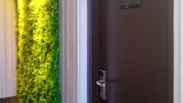 A hallway with a green vertical garden wall, a dark wooden door labeled '102', and a wooden floor with orange lines. - The Loft Plaza Hotel (Kajang)