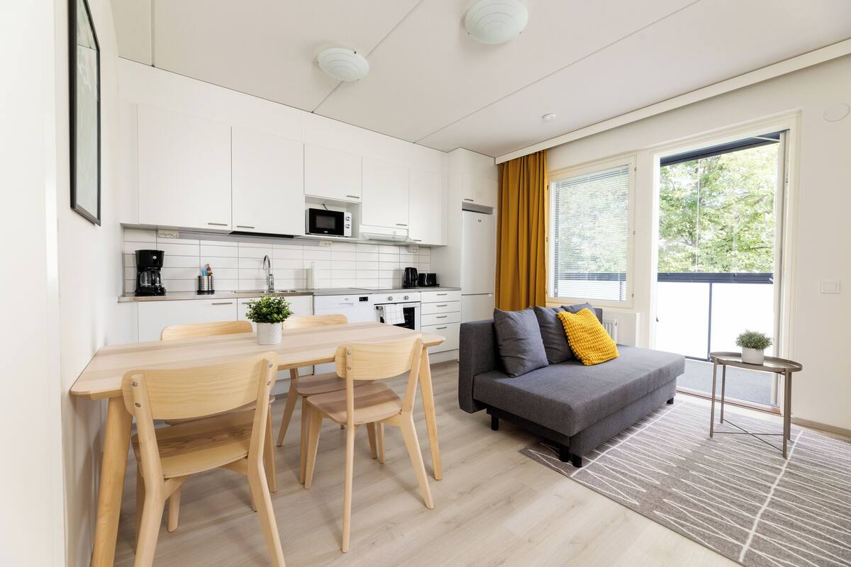 A modern kitchen with white cabinets, a dining table with chairs, a gray sofa with a yellow pillow, and a small side table with a plant.