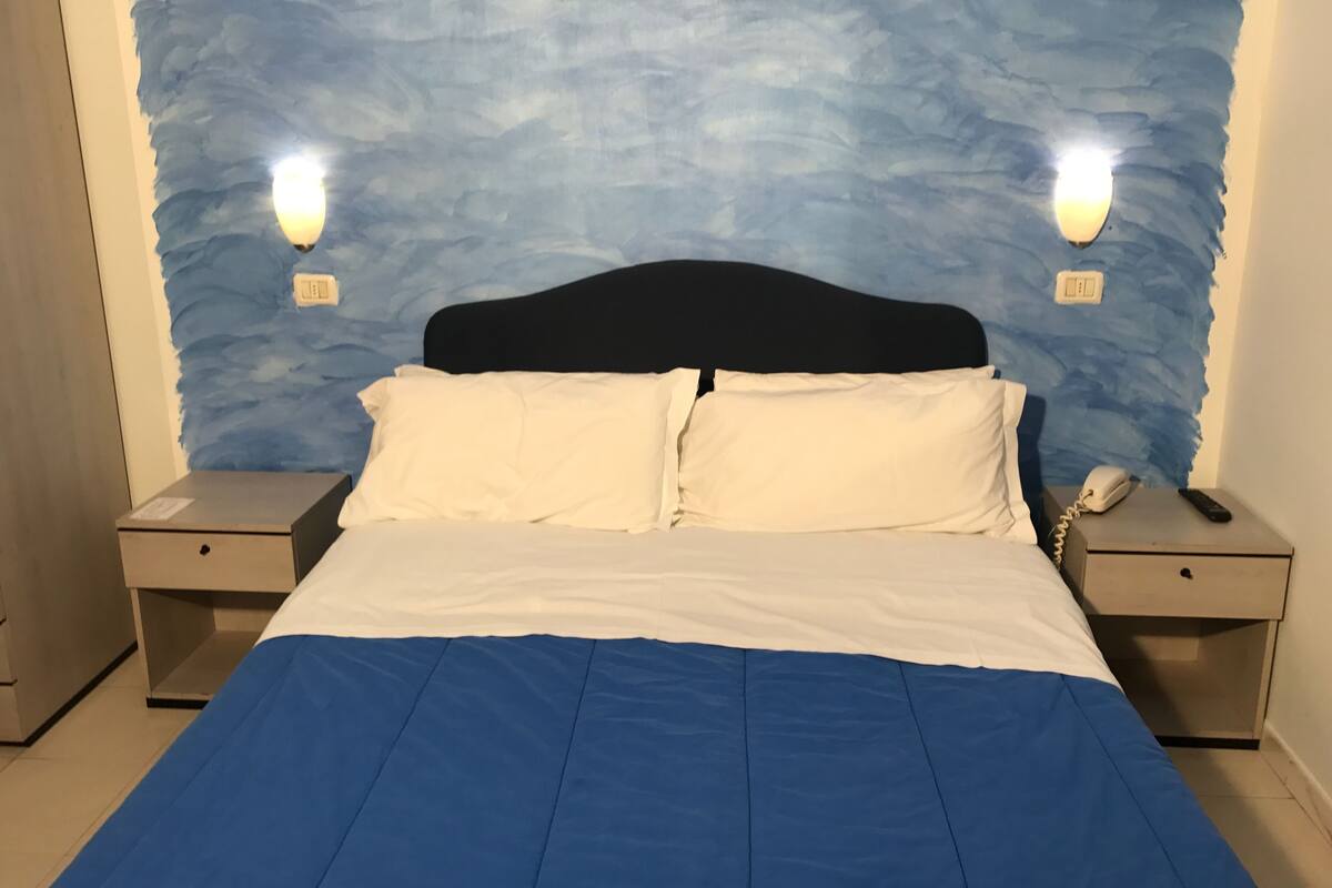 Una cama con ropa de cama blanca y azul, dos mesitas de noche con lámparas, y una pared azul con textura detrás de la cama.