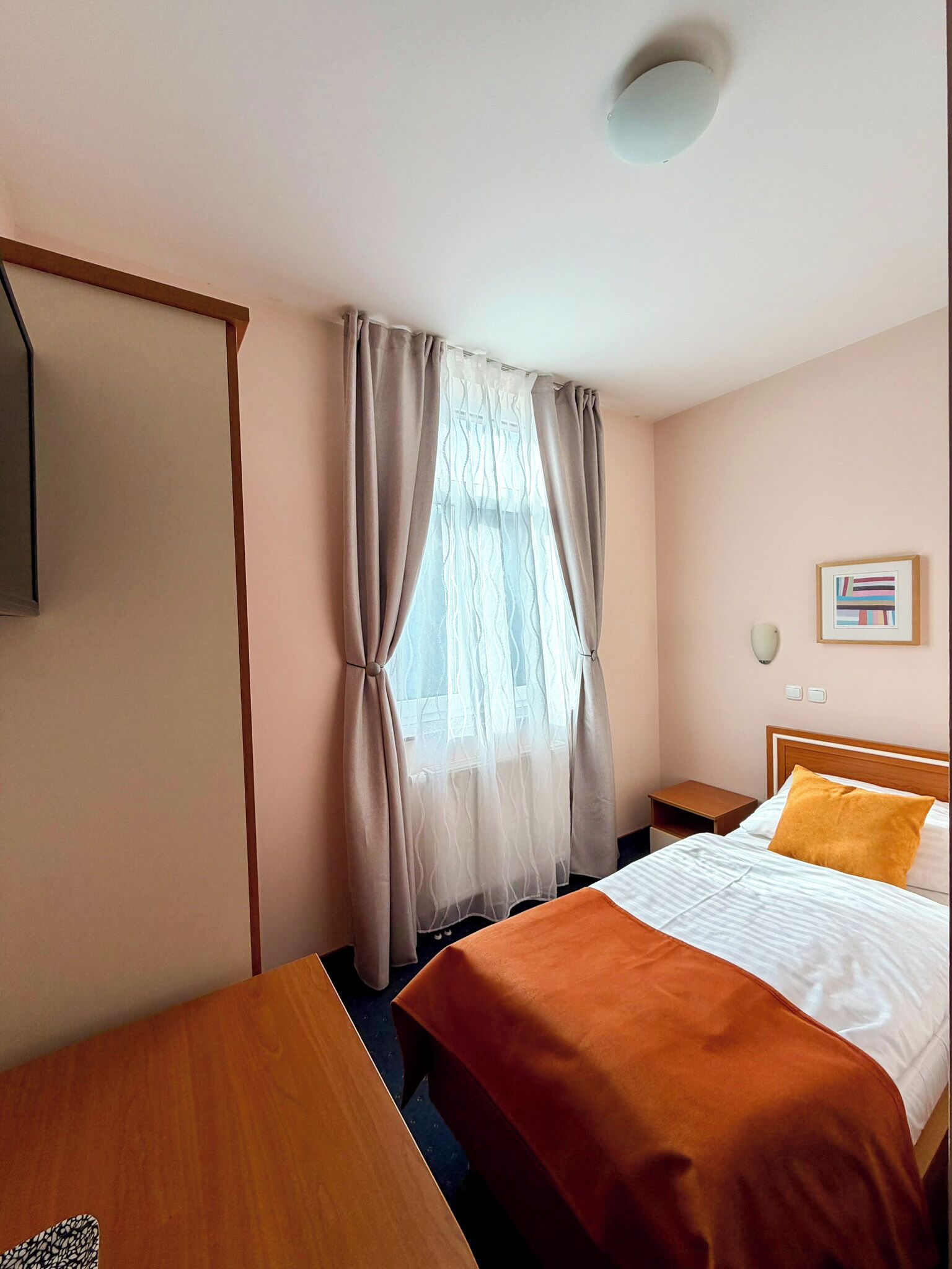 Hotellihuone, jossa on sänky, puinen yöpöytä, seinälle asennettu televisio ja ikkuna, jossa on verhot.