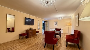A hotel room with a wooden floor, a television, a mirror, a red chair, a wooden cabinet, and a chandelier.