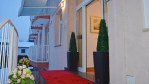 A hotel entrance with a red carpet, a balcony with white railings, and two potted plants. - Hotel Imperium (Moravske Toplice)