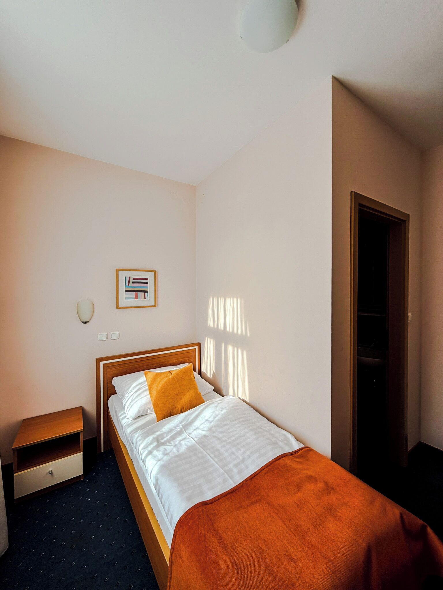 Habitación de hotel con cama, mesita de noche, un cuadro en la pared y una puerta a la derecha.
