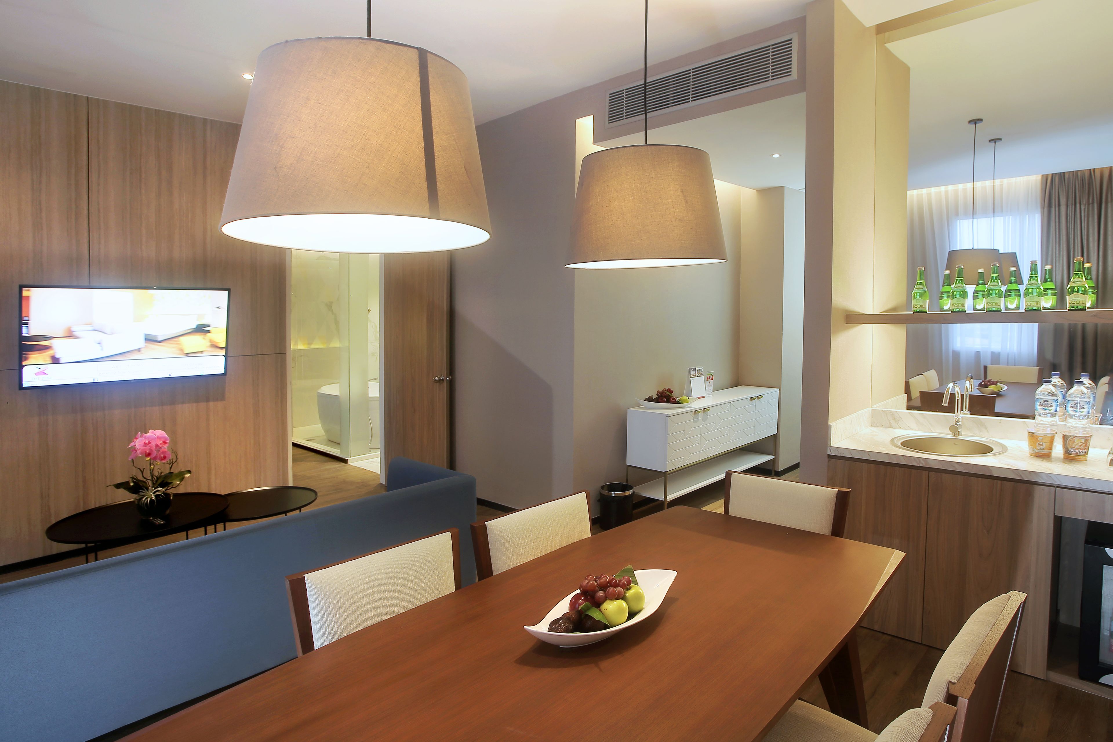 Une chambre d’hôtel moderne avec une table à manger en bois, un panier de fruits, un téléviseur à écran plat et un coin cuisine équipé d’un évier et d’un plan de travail.