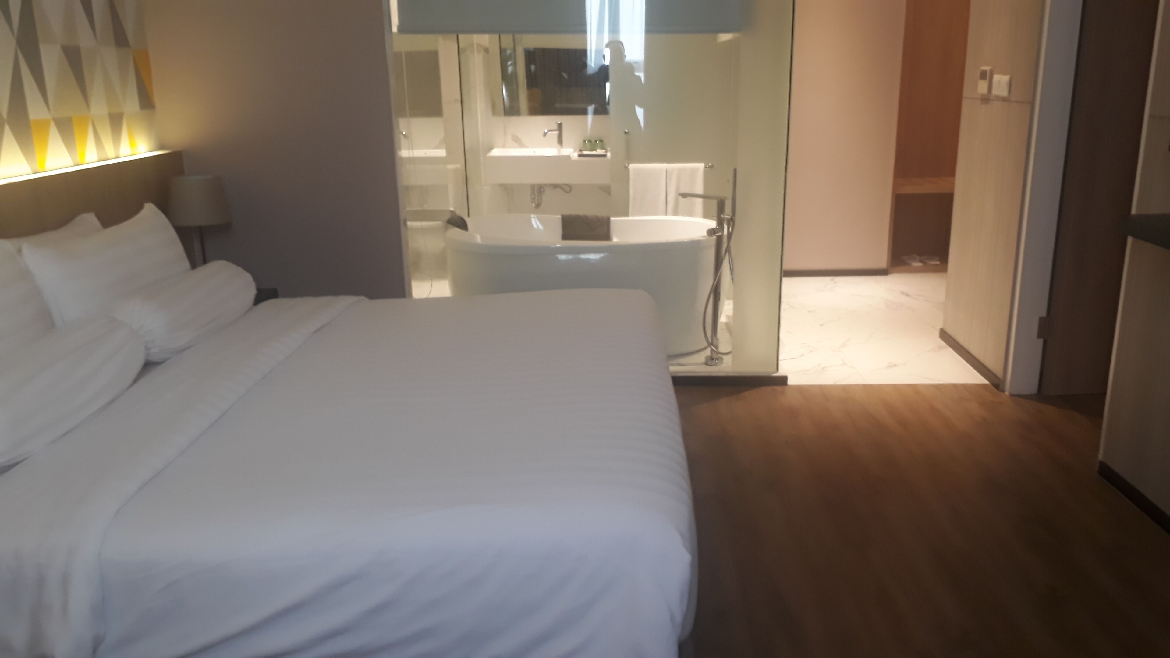 A hotel room with a large bed, a bathroom with a glass partition, and a wooden floor.