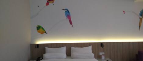 A hotel room with a bed, white pillows, a nightstand, and a wall with colorful bird murals.
