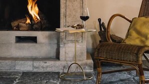 Una chimenea con fuego a la vista, una mesa con una copa de vino tinto y un sillón de mimbre.