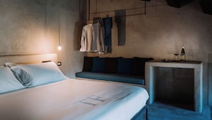 Un dormitorio con cama, un banco, una mesa con una botella y ropa colgada.