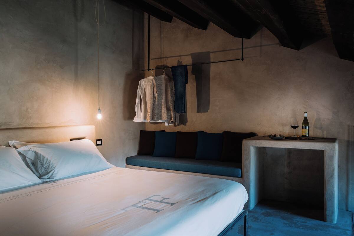 A bedroom with a bed, a bench, a table with a bottle, and hanging clothes.