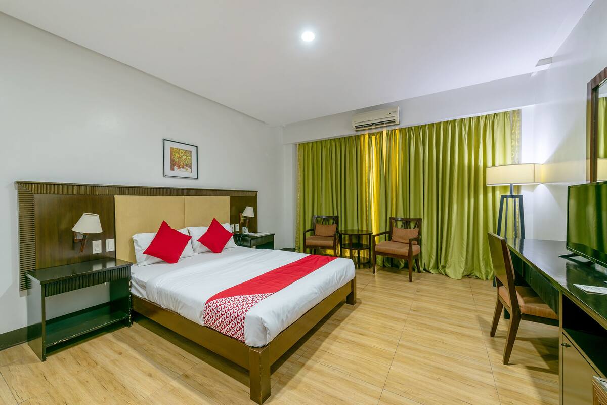A hotel room with a bed, a desk, a chair, a nightstand, and an air conditioning unit.