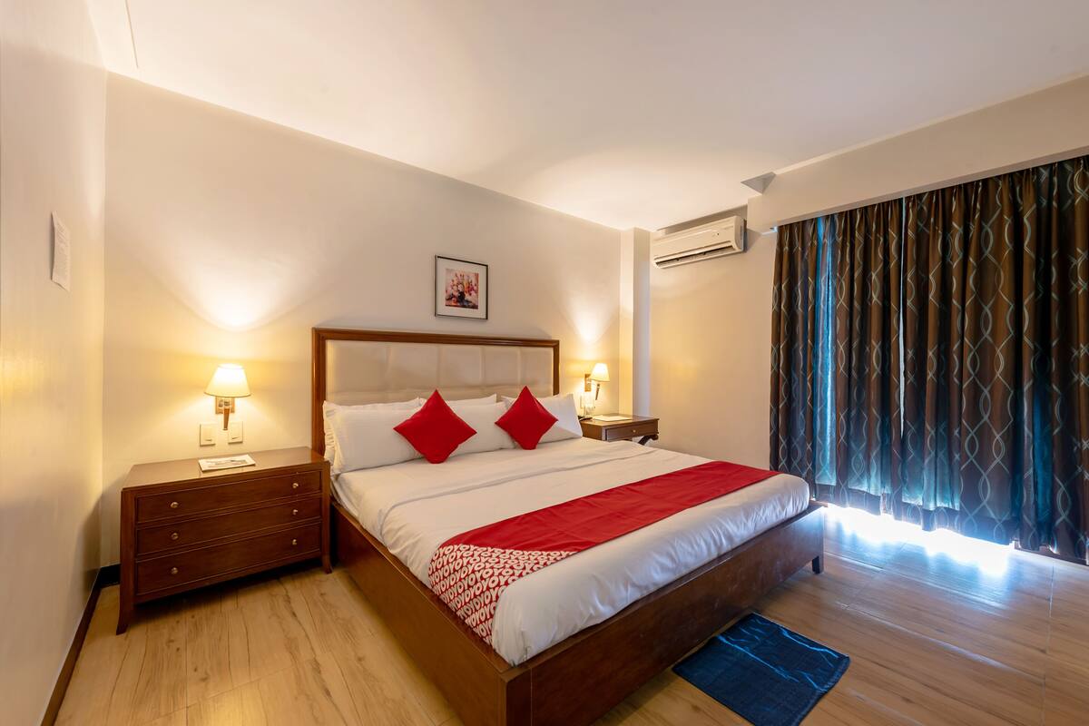 A hotel room with a large bed, wooden nightstands, a red throw blanket, and a view of the outdoors through curtains.
