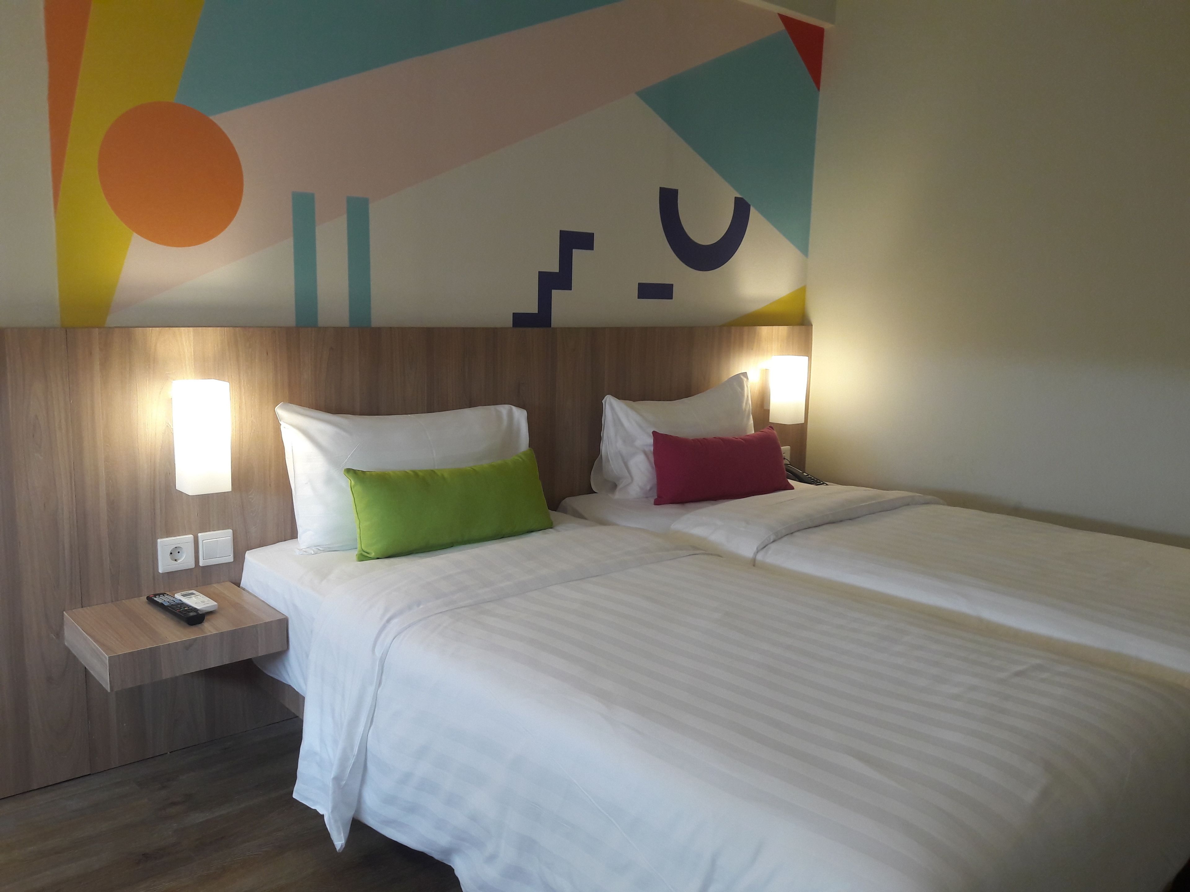 A hotel room with two beds, each with white bedding and colorful pillows. There are bedside tables with lamps and a remote control on one of them. The room has a wooden headboard and a colorful wall design.