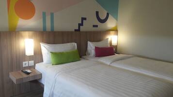 A hotel room with two beds, each with white bedding and colorful pillows. There are bedside tables with lamps and a remote control on one of them. The room has a wooden headboard and a colorful wall design.
