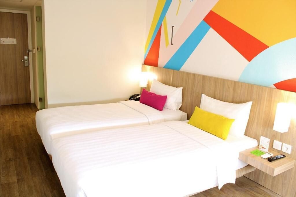 A hotel room with two beds, a wooden headboard, a nightstand with a lamp, and a colorful wall design.