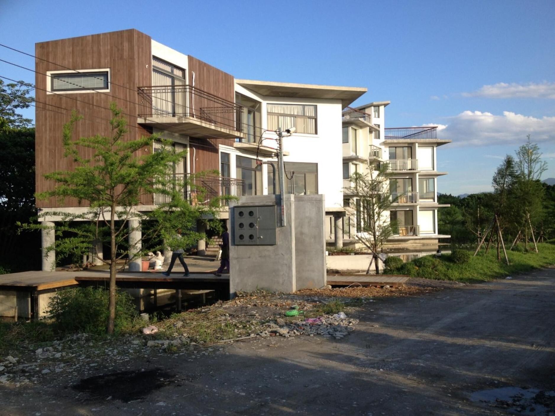 A modern multi-story residential building with balconies, a concrete utility box, and a paved driveway.